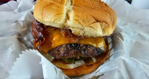 A close-up of a juicy cheeseburger with melted cheese, bacon, pickles, and lettuce on a soft bun, wrapped in paper.