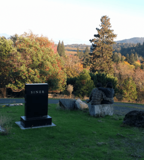 A couple sits on a rock, gazing at a scenic view near a black gravestone marked "Siner" surrounded by trees.