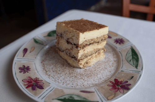 A slice of tiramisu layered dessert on a decorative plate, dusted with cocoa powder.