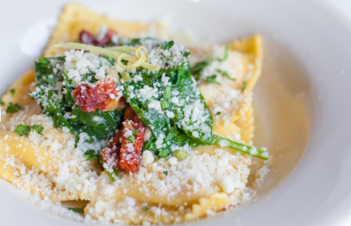 A plate of ravioli topped with spinach, sun-dried tomatoes, and grated cheese in a light sauce.