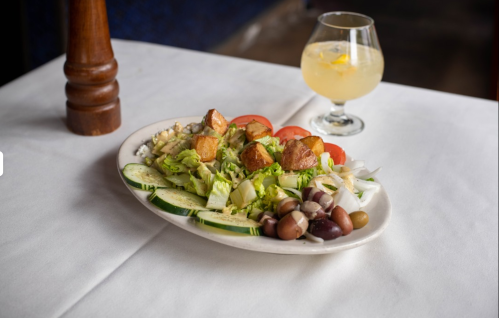 A fresh salad with cucumbers, tomatoes, croutons, and olives, alongside a glass of lemon drink on a white tablecloth.