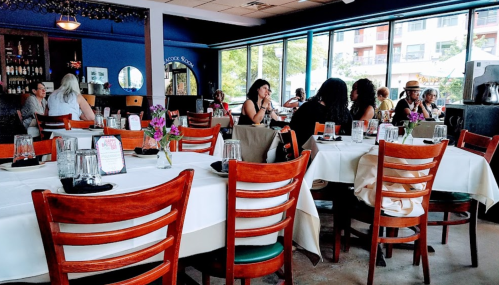A restaurant interior with tables set for dining, featuring patrons, flowers, and large windows letting in natural light.