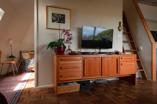 A cozy living room featuring a wooden TV stand, a television, and a decorative plant, with a staircase in the background.