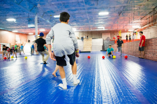 Children playing dodgeball in a spacious indoor gym with colorful balls and a blue floor.