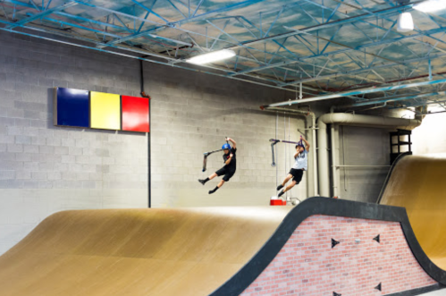 Two riders perform tricks on scooters in a skatepark with ramps and a colorful wall display in the background.