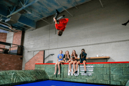 A person performs a backflip on a trampoline while four others watch from the side.