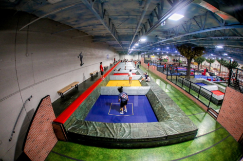 A spacious indoor parkour facility with multiple trampolines and training areas, filled with people practicing.