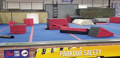 A parkour training area with various obstacles on a blue mat, featuring safety signage in the foreground.