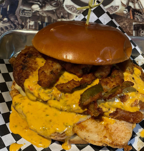 A stacked sandwich with fried chicken, jalapeños, and melted cheese on a toasted bun, served on a checkered paper.