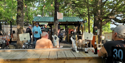 Outdoor music venue with people enjoying drinks, seated at tables under trees, and a band performing in the background.