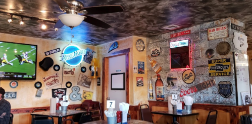 A cozy restaurant interior with vintage signs, a TV showing a game, and a ceiling fan. Tables are set for dining.