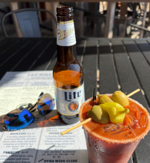 A close-up of a Bloody Mary with olives and a beer bottle on a table, with sunglasses and a menu in the background.