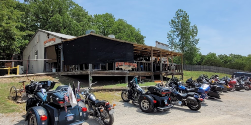 A rustic motorcycle bar with a large deck, surrounded by parked motorcycles on a sunny day.