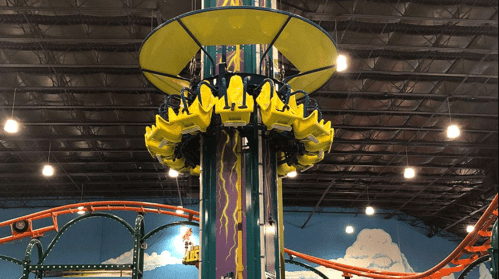A colorful amusement park ride with yellow seats, set against a blue sky mural and roller coaster tracks in the background.