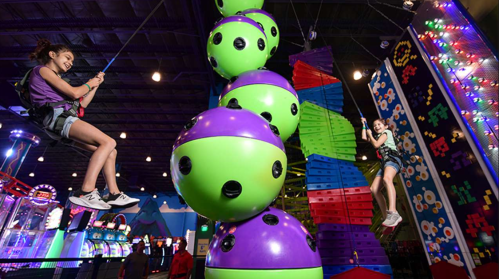 Two girls play on a colorful indoor climbing structure with large green and purple balls in a vibrant arcade setting.
