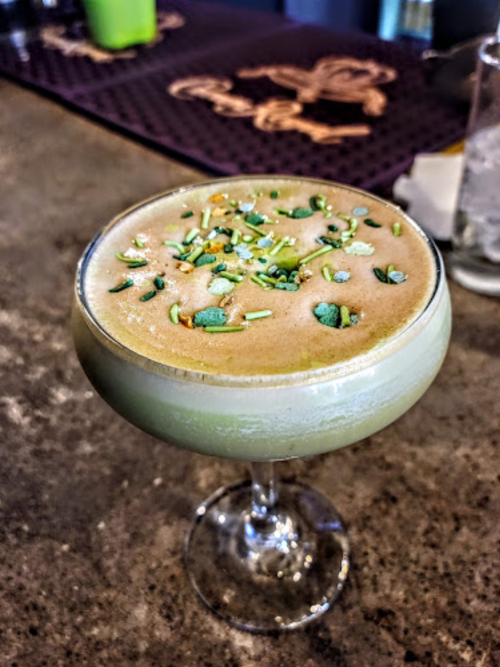 A frothy green cocktail garnished with herbs, served in a stylish glass on a bar countertop.