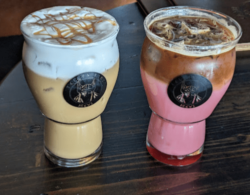 Two colorful drinks in glass cups: one with caramel topping and the other a pink iced beverage, both on a wooden table.