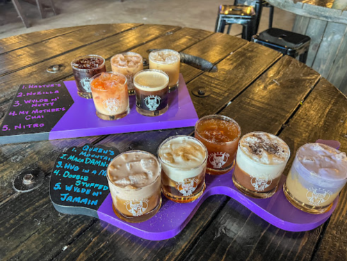 A wooden table with a flight of colorful craft beers and a chalkboard listing their names.