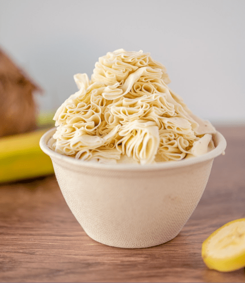 A bowl filled with intricately folded, pale yellow pasta, surrounded by a banana and a coconut in the background.