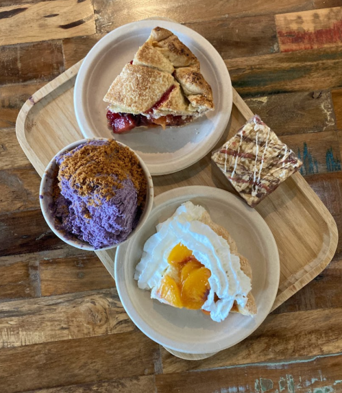 A wooden tray with a slice of pie, a bowl of purple ice cream, and a slice of cake topped with fruit and whipped cream.