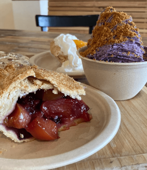 A slice of fruit pie with a flaky crust, a scoop of purple dessert, and a whipped cream-topped pastry on a wooden table.
