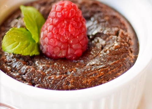 A rich chocolate dessert topped with a raspberry and a sprig of mint in a white ramekin.
