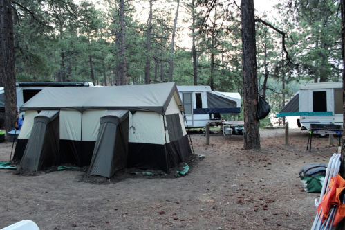 A camping site with a large tent and several trailers surrounded by tall trees and a dirt ground.