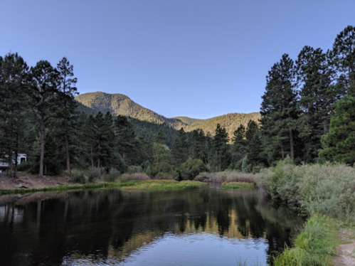 A serene river reflects mountains and trees under a clear blue sky, surrounded by lush greenery.