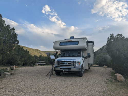 A parked RV on a gravel site surrounded by trees and mountains under a partly cloudy sky.