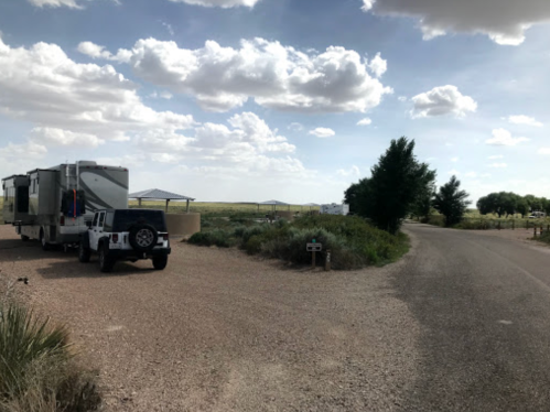 A gravel road leads to an RV park with parked RVs and a clear sky dotted with clouds. Green vegetation surrounds the area.
