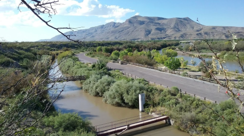 A scenic view of a winding river surrounded by greenery, with mountains in the background under a clear blue sky.