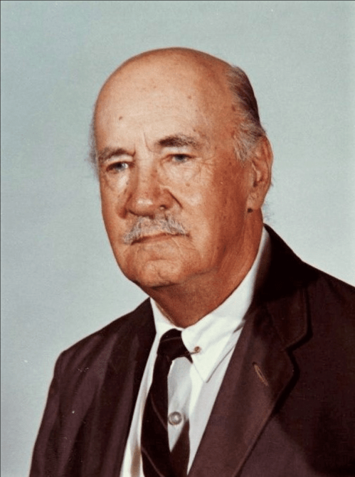 A middle-aged man with a bald head and mustache, wearing a suit and tie, looking serious against a plain background.