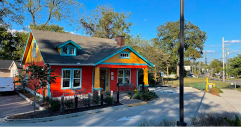 Colorful house with blue and orange exterior, surrounded by trees and a parking area, under a clear blue sky.