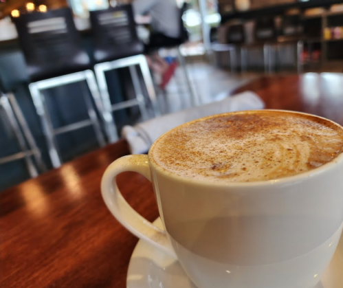 A close-up of a coffee cup with frothy milk, set on a wooden table in a cozy café. Background features blurred chairs.