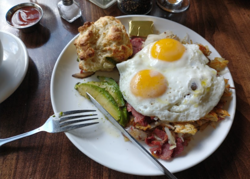 A plate of breakfast featuring two fried eggs, corned beef hash, avocado slices, a biscuit, and a pat of butter.