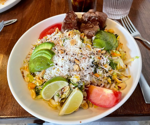 A colorful salad with shredded cabbage, tomatoes, avocado, peanuts, and two pieces of meat, served in a white bowl.