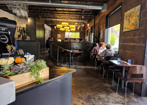 A cozy restaurant interior with wooden tables, plants, and patrons dining. Bright lighting and a welcoming atmosphere.