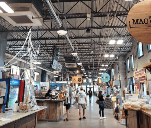A bustling market interior with vendors, shoppers, and various food stalls under a high ceiling.