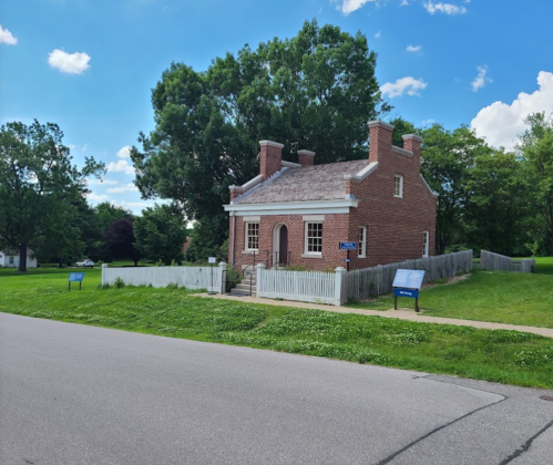 A historic brick house with a white picket fence, surrounded by green grass and trees, under a blue sky.