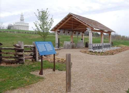 A gravel path leads to a pavilion with informational signs, surrounded by greenery and a historic building in the background.