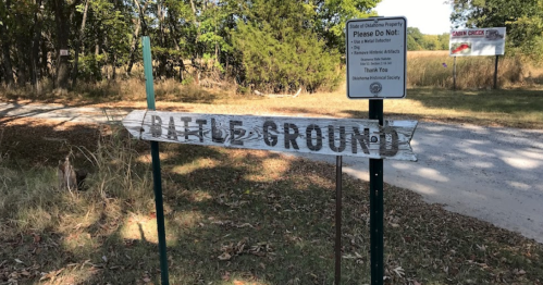 A weathered wooden sign reading "Battle Ground" near a dirt road, with a warning sign in the background.