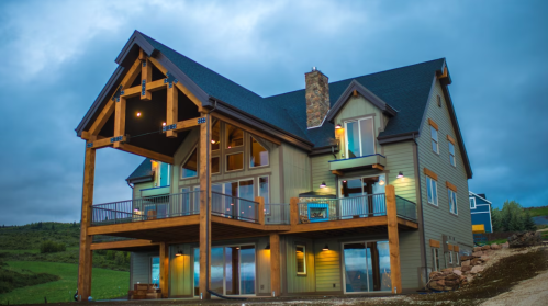 A large, modern mountain house with a wooden porch, large windows, and a stone chimney, set against a cloudy sky.
