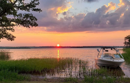 A serene sunset over a lake, with a boat anchored near the shore and lush greenery in the foreground.