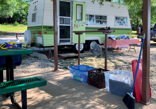 A vintage green and white camper parked under a shelter, with picnic tables and various items organized nearby.