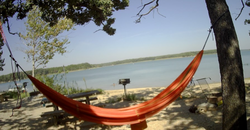 A vibrant orange hammock hangs between trees by a serene lake, with picnic tables and a grill nearby.