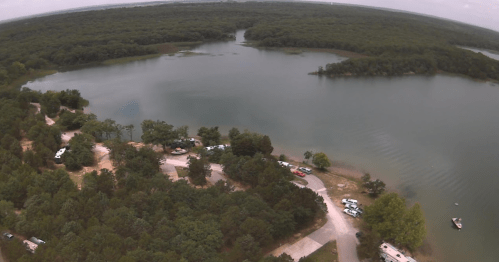 Aerial view of a serene lake surrounded by lush greenery and a winding road with parked vehicles.