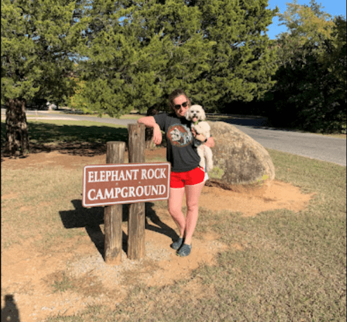 A person in a t-shirt and shorts stands by a sign for Elephant Rock Campground, holding a small dog.