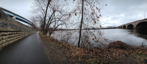 A cloudy riverside path with bare trees, a bridge overhead, and a stone wall along the water's edge.
