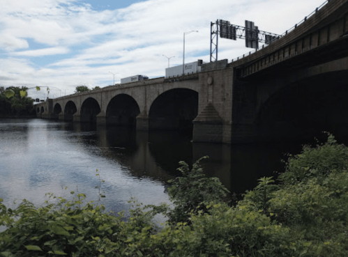 A bridge with arches spans a river, reflecting the sky and surrounded by greenery under a partly cloudy sky.