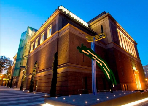 Modern building with a brick facade, illuminated at dusk, featuring a colorful abstract sculpture in the foreground.